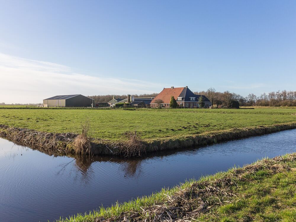 Brede sloot met spiegelend water langs groene weilanden bij Woudfennen aan de rand van Joure, met boerderij aan de horizon